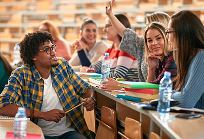 A group of people are sitting in a classroom, engaged in a discussion. There are several bottles of water on the table, and a book is open in front of one of the individuals. The classroom is furnished with chairs and a dining table. AI generated content
